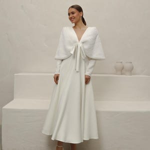 May include: A white wedding dress with a long-sleeve bodice and a flowing skirt. A matching faux fur shawl with a bow detail is worn over the shoulders. The model is wearing white heels. The background is a neutral color.