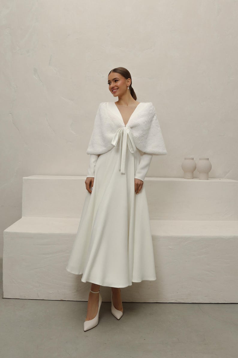May include: A white wedding dress with a long-sleeve bodice and a flowing skirt. A matching faux fur shawl with a bow detail is worn over the shoulders. The model is wearing white heels. The background is a neutral color.