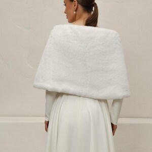 May include: A white faux fur capelet, likely for a wedding or formal event. The capelet is draped over a long-sleeved white dress. The image is taken from behind, showing the back of the person wearing the capelet and dress.