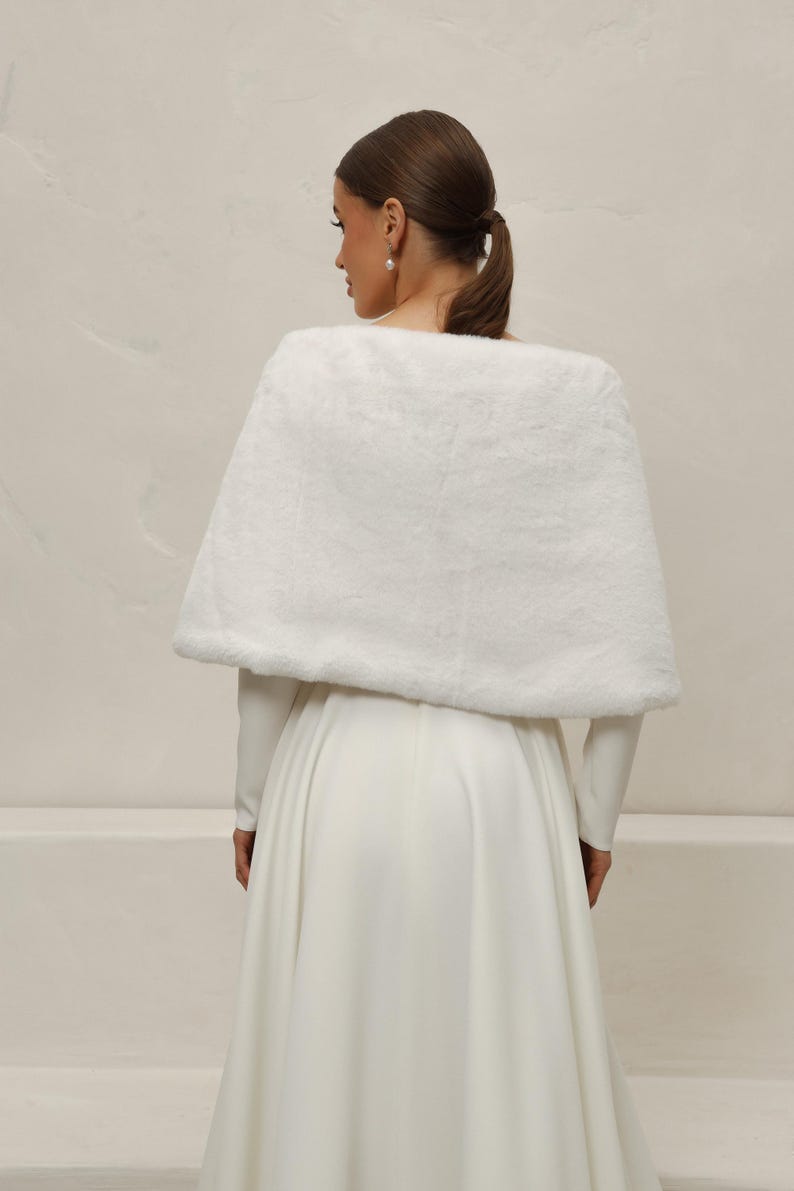 May include: A white faux fur capelet, likely for a wedding or formal event. The capelet is draped over a long-sleeved white dress. The image is taken from behind, showing the back of the person wearing the capelet and dress.