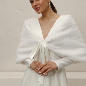May include: A white faux fur shawl with a satin tie closure. The shawl is draped over a white dress with long sleeves. The overall look is elegant and suitable for special occasions.