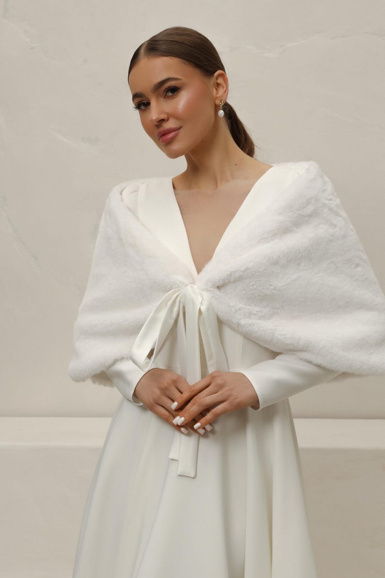 May include: A white faux fur shawl with a satin tie closure. The shawl is draped over a white dress with long sleeves. The overall look is elegant and suitable for special occasions.