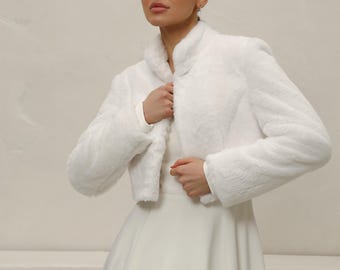 May include: A white faux fur bolero jacket with a stand-up collar. The cropped jacket is worn over a white dress. The image is set against a neutral background, highlighting the texture and style of the outerwear.