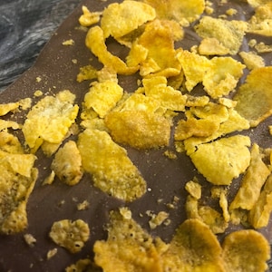 May include: A close-up shot of a chocolate bar topped with golden corn flakes. The chocolate is a rich brown, and the flakes are a bright yellow. The image highlights the textures of the chocolate and the cereal, creating a visually appealing treat.