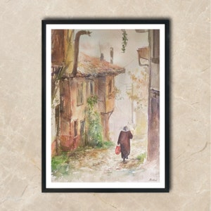 May include: Watercolor painting of a European street scene. A person with an umbrella walks down a cobblestone street past old buildings. The buildings are brown and beige with red tile roofs. The painting is framed in a black frame.