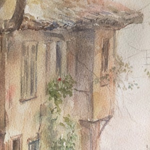May include: Watercolor painting of a weathered building with a tiled roof and small windows. The building's facade is a warm beige, with hints of pink and brown. Green foliage and a red flower add a touch of color to the scene.