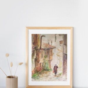 May include: Watercolor painting of a European street scene in a wooden frame. The artwork depicts a person walking down a cobblestone street lined with old buildings. The color palette includes browns, reds, and greens, creating a warm, inviting atmosphere.