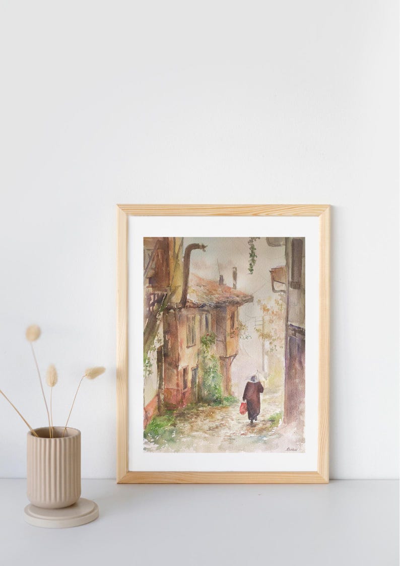 May include: Watercolor painting of a European street scene in a wooden frame. The artwork depicts a person walking down a cobblestone street lined with old buildings. The color palette includes browns, reds, and greens, creating a warm, inviting atmosphere.