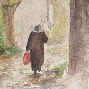 May include: Watercolor painting of a person walking in the rain under a clear umbrella. The person is wearing a dark coat and carrying a red bag. The scene is set on a cobblestone street with buildings and trees in the background.