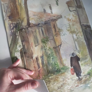 May include: Watercolor painting of a European street scene in muted tones. A person with an umbrella walks down a cobblestone street past a weathered building. The artwork has a vintage, atmospheric quality.