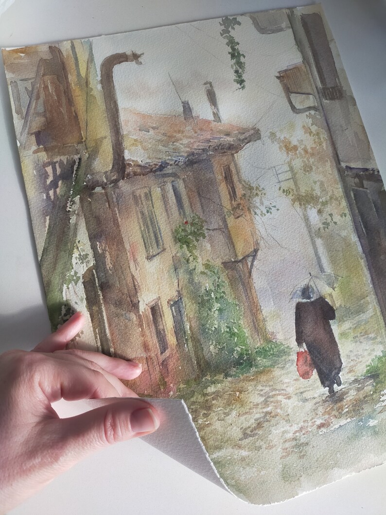 May include: Watercolor painting of a European street scene in muted tones. A person with an umbrella walks down a cobblestone street past a weathered building. The artwork has a vintage, atmospheric quality.