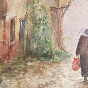 May include: Watercolor painting of a European street scene. A person with an umbrella walks down a cobblestone street, carrying a red bag. Buildings with windows and green foliage are on the left. The painting has a muted color palette.