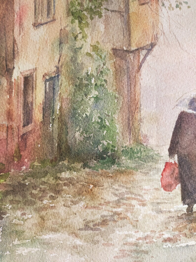 May include: Watercolor painting of a European street scene. A person with an umbrella walks down a cobblestone street, carrying a red bag. Buildings with windows and green foliage are on the left. The painting has a muted color palette.