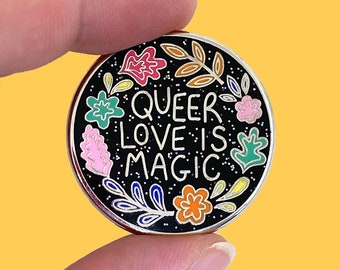 May include: A round enamel pin with a black background and the words "QUEER LOVE IS MAGIC" in white. The pin is decorated with colorful floral and leaf designs in pink, orange, green, and blue. The pin has a silver border.
