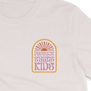 以下が含まれることがあります： レトロなスタイルのグラフィックデザインが施された白いTシャツ。グラフィックには、光線を放つ太陽、アーチ、ピンクとオレンジの「Protect Trans Kids」というテキストが描かれています。