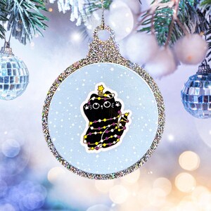 May include: A silver glitter Christmas bauble featuring a black cat adorned with a string of yellow and purple lights. The cat is wearing a star on its head. The bauble has a blue background with white snowflakes.