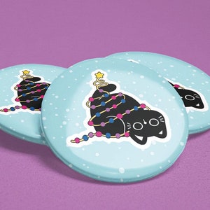 May include: Three blue and white round buttons featuring a black cat entangled in Christmas lights. The cat has a festive expression and a star on top of its head.