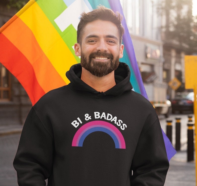 May include: A black hoodie with a rainbow graphic and the text "Bi & Badass". The person wearing the hoodie is standing in front of a rainbow pride flag.