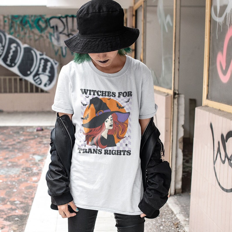 Goth person wearing a White crew neck shirt featuring the slogan Witches for Trans Rights with an illustrated graphic of a witch with bats flying around her hat