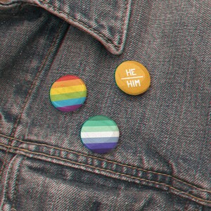 Puede incluir: Tres pines de bot&oacute;n en una chaqueta vaquera. Un pin es de color arco&iacute;ris, uno es azul y verde, y uno es amarillo con el texto "HE/HIM" en blanco.