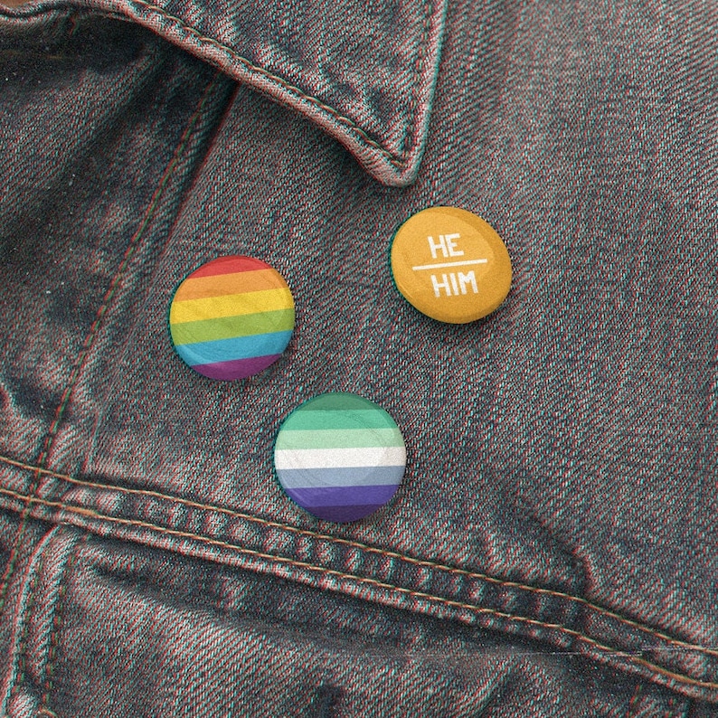 Puede incluir: Tres pines de bot&oacute;n en una chaqueta vaquera. Un pin es de color arco&iacute;ris, uno es azul y verde, y uno es amarillo con el texto "HE/HIM" en blanco.