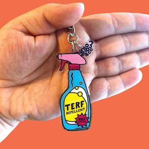 May include: A novelty keychain with a blue and yellow spray bottle design. The keychain reads "TERF REPELLENT" and "100% EFFECTIVE". It has a pink spray nozzle and a silver chain.