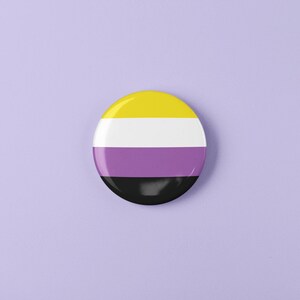 May include: A round button with the colors of the non-binary pride flag. The button is yellow, white, purple, and black.