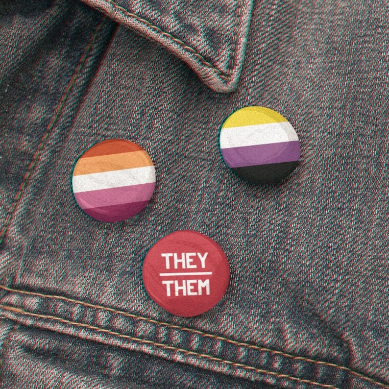 May include: Three button pins on a denim jacket. The first pin is a lesbian pride flag, the second is a non-binary pride flag, and the third is a red pin with the text "They/Them".