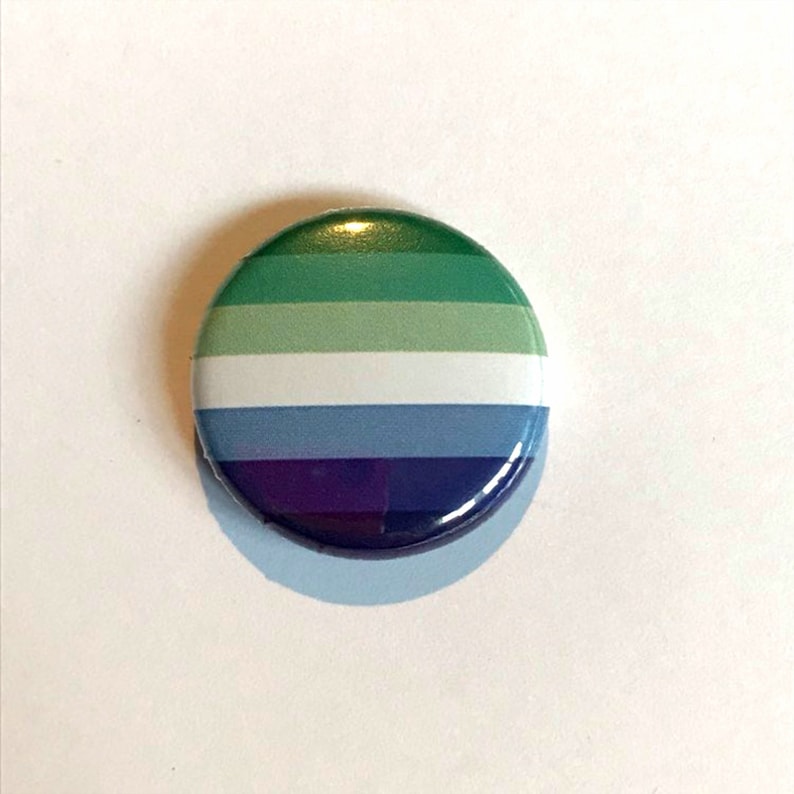 Puede incluir: Un bot&oacute;n redondo con un dise&ntilde;o a rayas verde, verde claro, blanco, azul y morado. El bot&oacute;n es una representaci&oacute;n de la bandera del orgullo genderqueer.