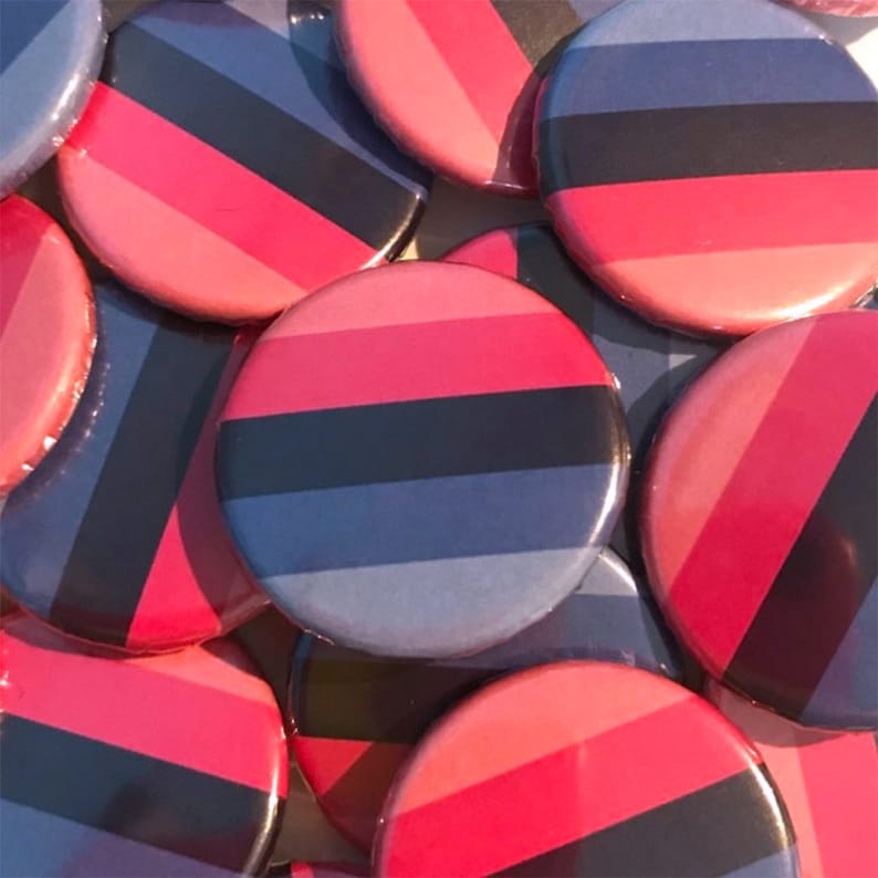 May include: A close-up of several round buttons with a black, pink, and blue striped design. The buttons are arranged in a pile.