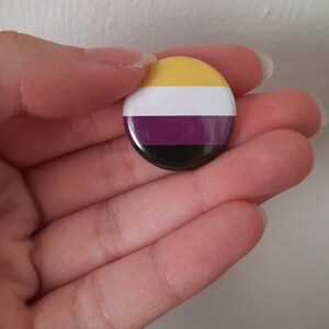 May include: A round button with the colors of the non-binary pride flag. The flag has yellow, white, purple, and black stripes.