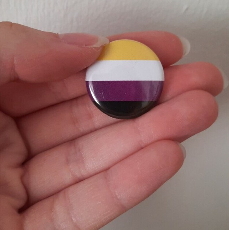 May include: A round button with the colors of the non-binary pride flag. The flag has yellow, white, purple, and black stripes.