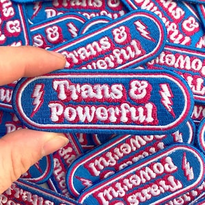 May include: A blue and pink embroidered patch with a lightning bolt and the text "Trans & Powerful".