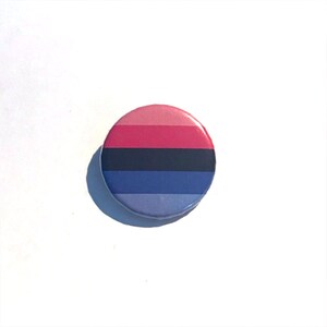 May include: A round button with a pink, black, blue, and purple striped design. The button is a representation of the bisexual pride flag.