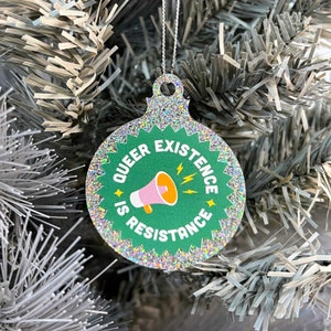 Peut inclure: Une boule de Noël verte et argentée à paillettes avec le texte "Queer Existence is Resistance" et un mégaphone rose avec un éclair jaune.