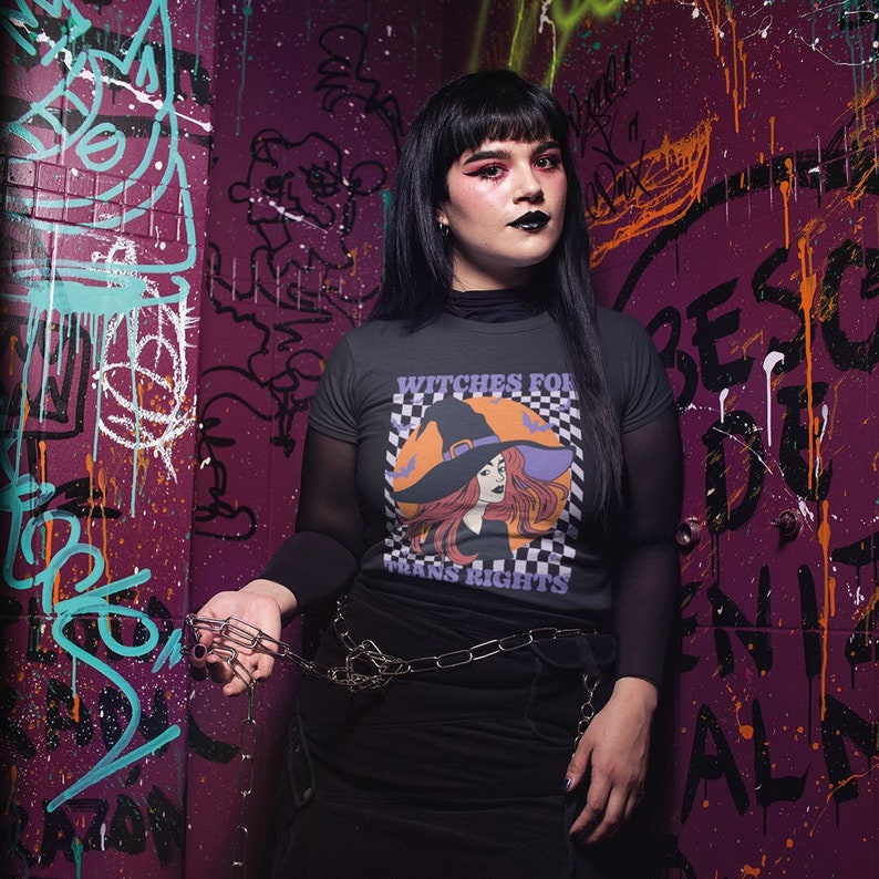 Goth person wearing a Dark Heather Grey crew neck shirt featuring the slogan Witches for Trans Rights with an illustrated graphic of a witch with bats flying around her hat