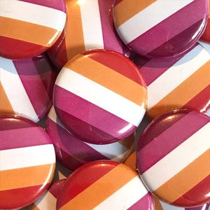 May include: A close-up of several round buttons with the lesbian pride flag colors: orange, white, pink, and red. The buttons are arranged in a pile, with some overlapping.