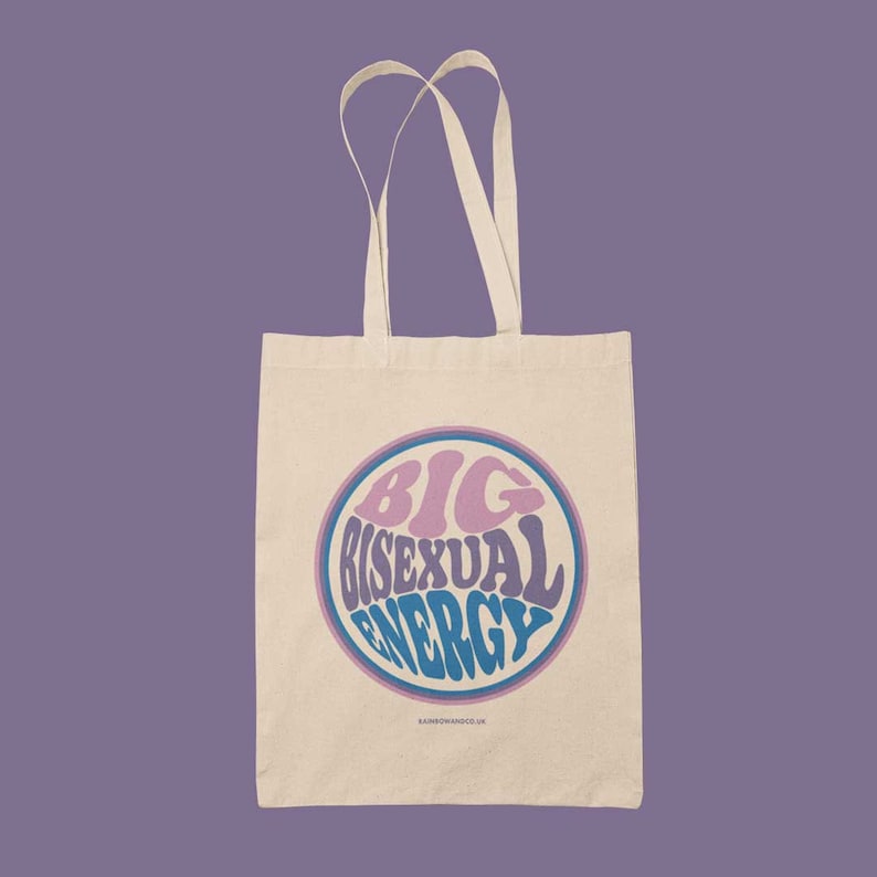 K&ouml;nnte beinhalten: Eine beige Tragetasche mit einem blauen und rosa Kreisdesign, auf dem in einer Retro-Schrift "BIG BISEXUAL ENERGY" steht.