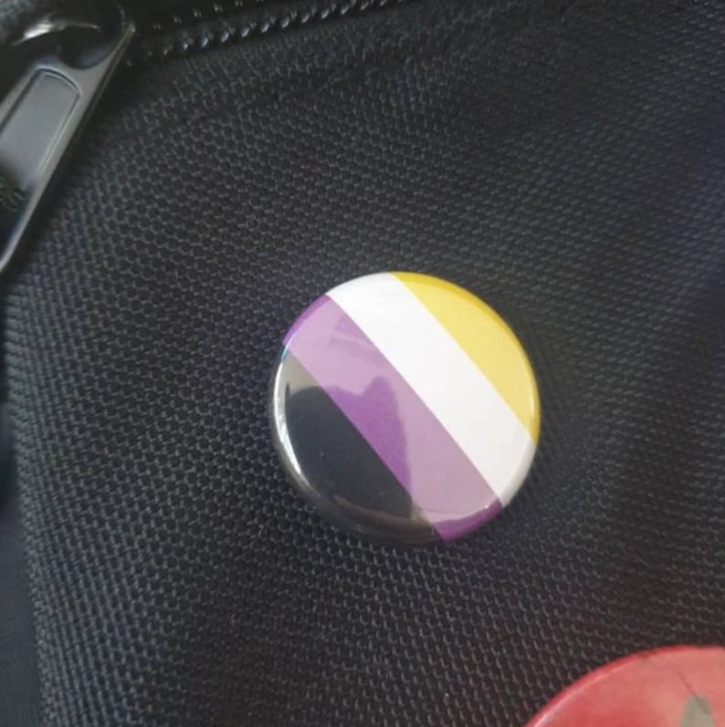 May include: A round button with the colors of the non-binary pride flag. The button is black, purple, white, yellow, and has a shiny finish.
