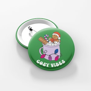 May include: Green button with a purple mug filled with marshmallows, chocolate, and a gingerbread man. The mug is decorated with a string of lights and the text "Cosy Vibes".