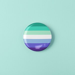 Puede incluir: Un bot&oacute;n redondo con un dise&ntilde;o a rayas en tonos de verde, azul y morado. El bot&oacute;n representa la bandera del orgullo genderqueer.