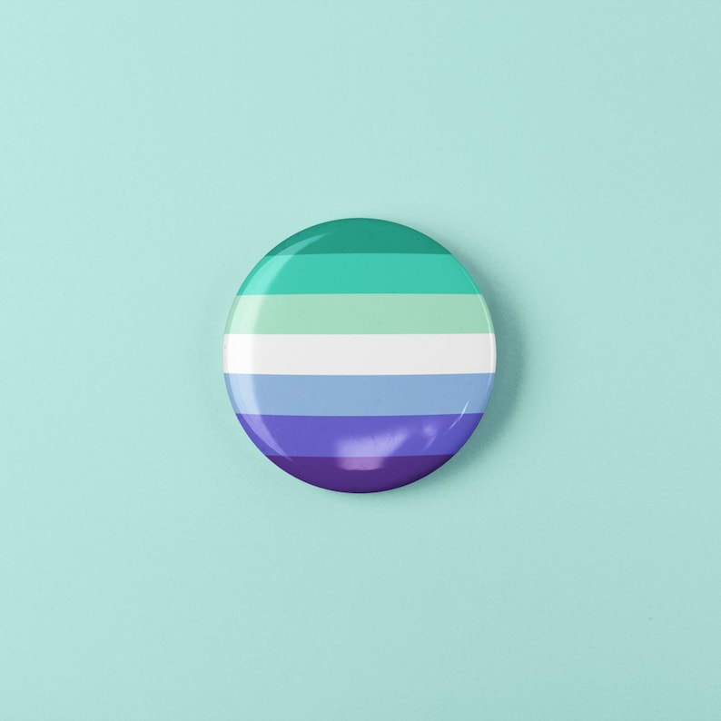 Puede incluir: Un bot&oacute;n redondo con un dise&ntilde;o a rayas en tonos de verde, azul y morado. El bot&oacute;n representa la bandera del orgullo genderqueer.