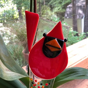 May include: A red glass cardinal bird plant stake with black sunglasses and an orange beak. The plant stake is in a turquoise ceramic pot with an orchid plant. The cardinal has a clear glass body with colorful dots.