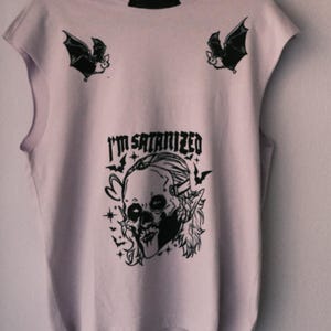 Op de afbeelding: Een lichtroze mouwloos T-shirt met een zwarte grafische print. Het ontwerp bevat twee vleermuizen bij de schouders en een centrale illustratie van een schedel met lang haar, omringd door kleine vleermuizen en sterren, met de tekst I'M SATANIZED.