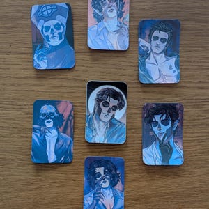 May include: A collection of seven rectangular stickers featuring illustrations of a band member with theatrical makeup. The stickers have a blue and white color scheme, with the band member in various poses and outfits.