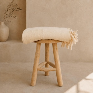 May include: A beige woven throw blanket with a subtle diamond pattern and knotted fringe edges draped over a light-coloured wooden stool with three legs and a central support structure, featuring a textured surface and a neutral colour palette.