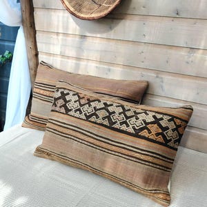 Peut inclure: Deux coussins rectangulaires avec un motif tissé. Les coussins présentent une palette de couleurs marron et beige avec des motifs géométriques et rayés. Les coussins sont placés sur une surface blanche contre un mur en bois.