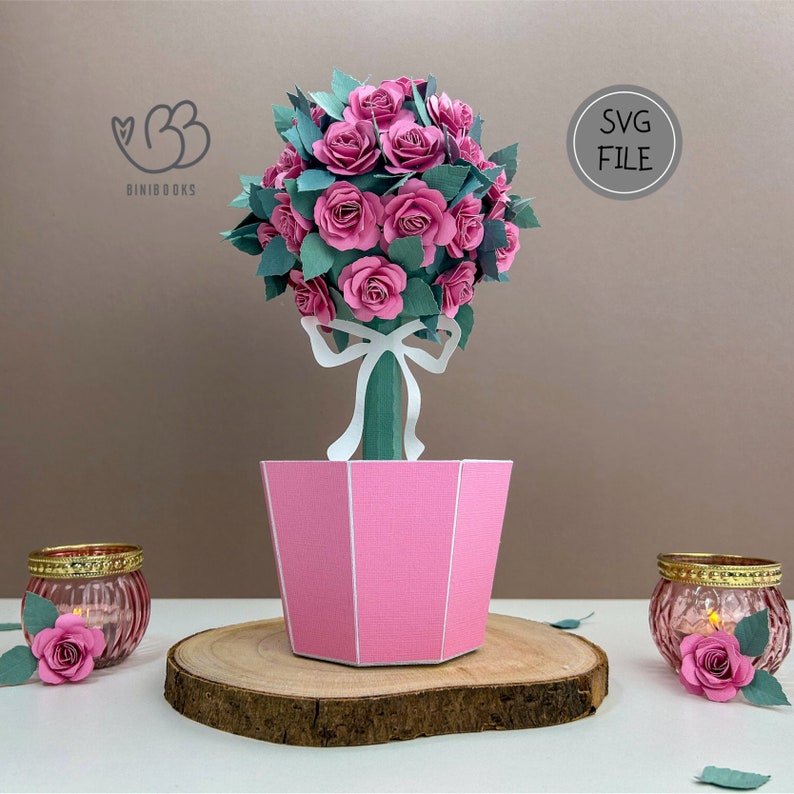 May include: A pink papercraft rose topiary with a white bow and green leaves in a pink pot. The topiary is sitting on a wooden slice. The image also includes the text "SVG FILE".