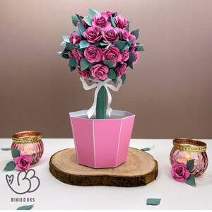 May include: A pink papercraft rose topiary with a white ribbon and green leaves in a pink pot. The topiary is sitting on a wooden base.