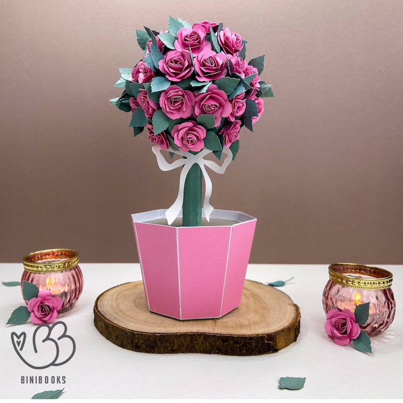 May include: A pink papercraft rose topiary with a white ribbon and green leaves in a pink pot. The topiary is sitting on a wooden base.